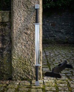 Double ended viking sword with a Celtic knotwork pommel at each end and twin cord wrapped grips standing against a granite pillar on mossy cobblestones