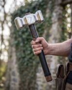 Double faced steel war hammer with tooled black leather grip held in one hand in front of an ivy covered wall