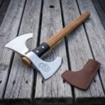 Double headed steel axe with etched coiled serpent and oak haft lying on grey wooden boards beside a brown leather head cover