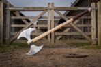 Double headed steel Viking axe with twin crescent bits and etched beast standing on its ash haft before a wooden farm gate