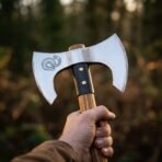 Double headed Viking axe with mirror polished blades and serpent etching held up by the oak haft against autumn woods
