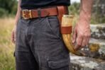 Drinking horn with brass rim carried at the hip in a tan leather belt hanger with brass buckle on a man in grey trousers