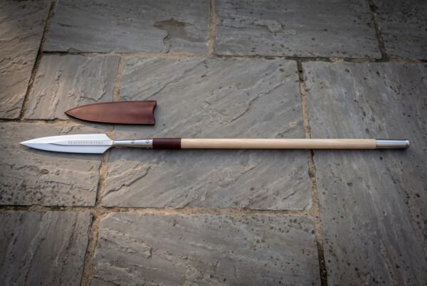 Elder Futhark spear with a polished leaf shaped head and a rune etched ash haft standing against a dry stone wall in low winter sun