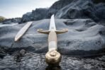 End on view of a steel sabre with rolled brass pommel, ribbed bone toned grip and scrolled quillons on a wet river stone
