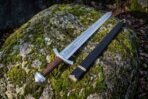 Engraved high carbon steel Viking sword with cord wrapped grip lying on a mossy boulder beside its black scabbard