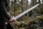 Engraved high carbon steel Viking sword with downswept cross held out in one hand in a rocky forest