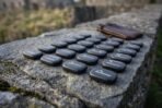 Close up of engraved slate rune stones laid out on a mossy stone wall with a brown leather drawstring pouch and ruins behind