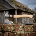 Engraved valknut axe with a damascus pattern welded head, hammer poll and curved ash haft resting on a lichen covered brick wall by a turf longhouse