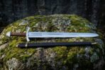 Engraved viking sword lying on a moss covered boulder with a cut vine and beast panel, cord wrapped grip and black scabbard with brass throat and chape