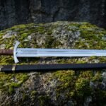 Engraved viking sword lying on a moss covered boulder with a cut vine and beast panel, cord wrapped grip and black scabbard with brass throat and chape