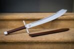Falchion with curved fullered blade, steel cross guard and brown leather grip on a sandstone step beside its scabbard
