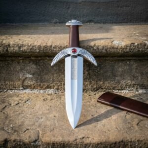 Fantasy dagger with a winged steel guard, red cabochon stone and broad leaf blade standing on a weathered sandstone step beside its leather scabbard