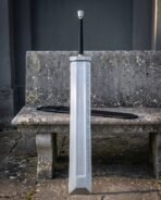 Fantasy greatsword with a broad slab sided blade and two rune etched lines standing against a weathered stone bench beside a long black leather scabbard