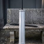 Fantasy greatsword with a broad slab sided blade and two rune etched lines standing against a weathered stone bench beside a long black leather scabbard