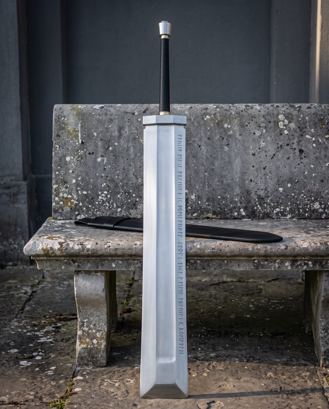 Fantasy greatsword with a broad slab sided blade and two rune etched lines standing against a weathered stone bench besi Fantasy greatsword with a broad slab sided blade and two rune etched lines standing against a weathered stone bench beside a long black leather scabbard