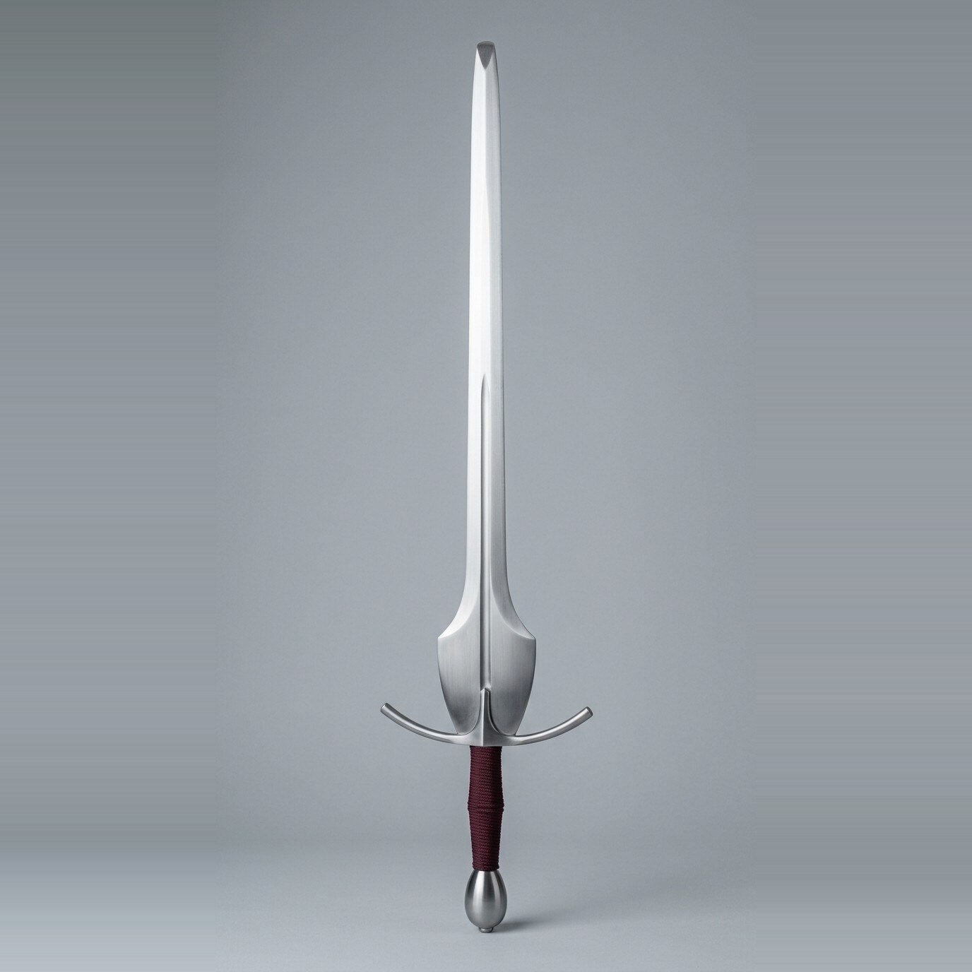 Brimhalt feder with shield schilt and lower fuller Feder training sword with a shield shaped schilt, curved crossguard, a narrow fuller low in the blade and a burgundy cord wrapped grip standing point up against a plain grey studio backdrop