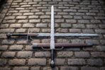 Flamberge longsword with an undulating flame bladed edge and black leather grip lying crossed with a second sword on cobblestones beside a brown leather scabbard