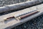 Flamberge sword with polished wavy blade, brass guard and red leather grip lying on driftwood beside its red brown scabbard