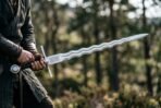 Flamberge sword with undulating blade held out in both hands by a man in a dark cloak among pine trees