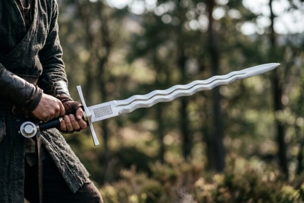 Flamberge sword with undulating blade held out in both hands by a man in a dark cloak among pine trees