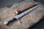 Flamberge sword with wavy blade, etched knotwork panel and disc pommel on a slate slab beside its wooden scabbard