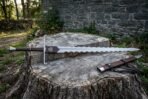 Flame bladed arming sword with scrolled crossguard and Valknut rain guard lying on a tree stump beside its leather scabbard