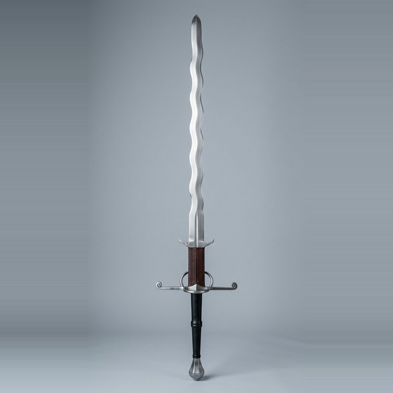 Vulkbrand flame bladed greatsword with parierhaken Flame bladed greatsword with waved edges on both sides, scrolled quillon tips, a side ring and a leather wrapped ricasso above the parierhaken laid on a plain grey studio backdrop