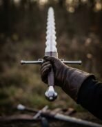Flame bladed longsword with a wavy steel blade, ricasso and leather grip held upright in a gloved hand in a dark forest