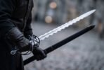 Flame bladed short sword with blackened swept ring hilt held in black gloves beside its black scabbard on a cobbled street