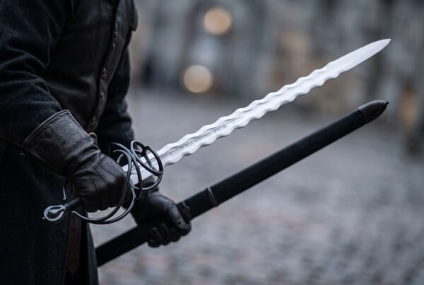 Flame bladed short sword with blackened swept ring hilt held in black gloves beside its black scabbard on a cobbled street