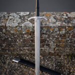 Fluted pommel damascus longsword standing against a lichened sandstone wall on gravel, its pattern welded blade, downswept guard and black leather scabbard fully in view