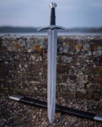 Fluted pommel damascus longsword upright on a weathered sandstone parapet at dusk, the wood grain pattern of the blade and the black scabbard lying on gravel below