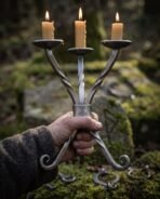 Forged twisted steel candle holder with three lit beeswax candles held up in one hand over a mossy stone