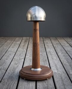 Four panel viking helmet in brushed steel displayed on a turned walnut stand carved with Elder Futhark runes, standing on grey timber decking