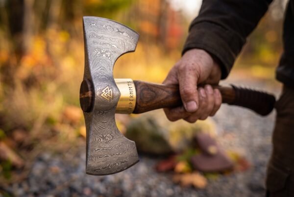 Front view of a Damascus double bit head with valknut inlay and engraved brass collar held by a hand on a woodland track