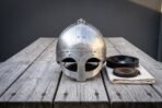 Front view of a riveted steel spectacle helmet with engraved nasal and eye guards next to a padded liner and chin strap