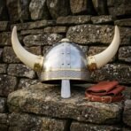 Front view of a riveted steel Viking helmet with pale curved horns and engraved browband on a mossy stone wall