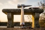 Front view of a steel war hammer with square face and long curved beak on a pale ash haft leaning on a stone bench at dusk