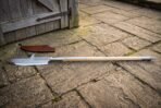 Full length glaive polearm with a pale ash haft and steel butt spike lying on flagstones by a wooden door