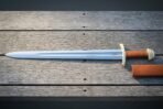 Full length Norse sword with fullered steel blade, etched raven and brass fittings lying on grey boards next to its tan scabbard