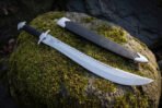 Full length polished sawback short sword beside its black leather sheath on a mossy boulder, showing the etched chevrons, compass roundel and forward swept steel guard.