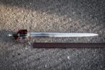 Full length steel basket hilt broadsword with red liner lying on gravel above its brown leather scabbard