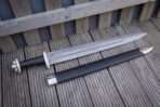 Full length view of a celtic knot panel viking sword beside its black leather scabbard on grey decking, showing the broad pattern welded blade and curved lower guard