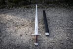 Full length view of a chevron fuller viking sword and its black leather scabbard laid on gravel, showing the broad fullered blade, brown grip and lobed steel pommel