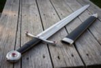 Full length view of a jewelled arming sword in 1095 steel on grey deck boards with the black leather scabbard alongside, red cabochons bright in the quillon tips
