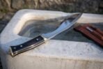 Full tang kukri knife with black buffalo horn handle angled across a stone trough, leather sheath and companion knives behind