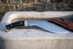 Full tang kukri with polished curved blade and buffalo horn grip on a stone trough, side view with leather sheath