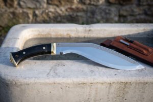 Full tang kukri with polished curved blade and buffalo horn grip on a stone trough, side view with leather sheath