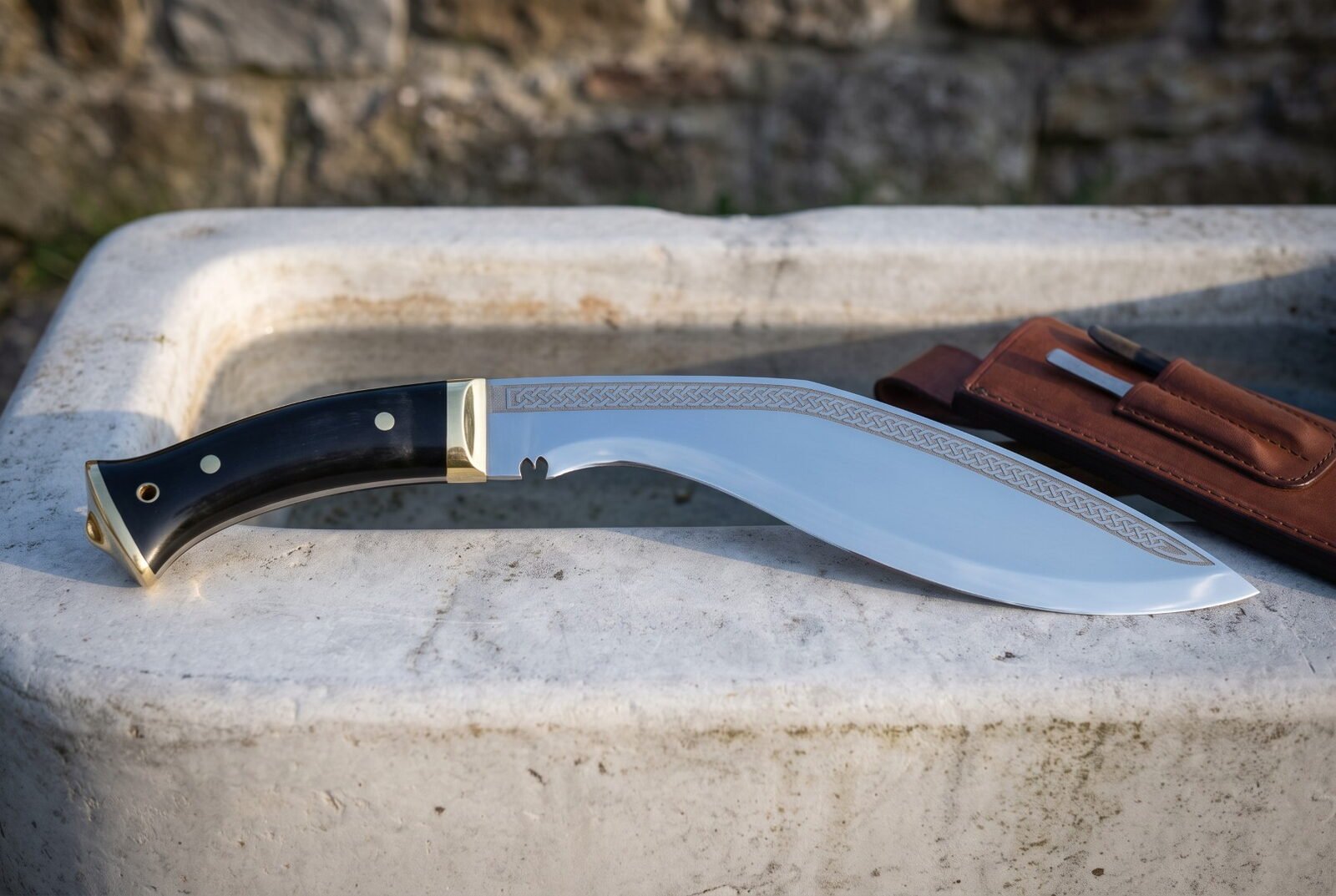 Full tang kukri with a polished blade and etched knotwork spine, black buffalo horn handle and brass fittings resting on a white stone trough beside a leather sheath