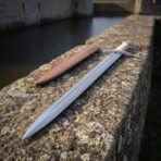 Fullered steel Celtic sword seen point first along a stone wall above a moat, tan leather scabbard beside it